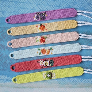 May include: Six colourful friendship bracelets with embroidered fruit and floral designs. The bracelets are in shades of purple, yellow, pink, light blue, red, and green. Each bracelet has a loop and white string for tying.