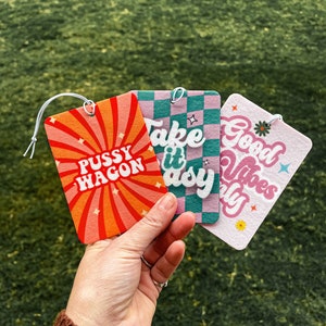 Könnte beinhalten: Drei Auto-Lufterfrischer mit verschiedenen Designs. Der erste ist orange und rot mit dem Text "Pussy Wagon". Der zweite ist rosa und grün mit dem Text "Take it Easy". Der dritte ist rosa und grün mit dem Text "Good Vibes Only".