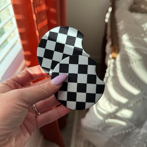 Puede incluir: Dos posavasos de coche a cuadros blancos y negros. Los posavasos son redondos con una pequeña muesca en un lado. El fondo incluye una cortina naranja y una cama con una colcha blanca.