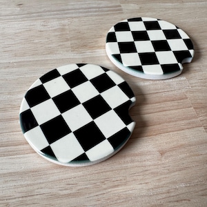 May include: Two circular car cup holder coasters featuring a black and white checkered pattern. Each coaster has a small semi-circular notch cut out from the edge, designed for easy removal from a vehicle cup holder console.