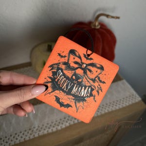 May include: Orange car air freshener with a black and white illustration of a spider web, bow, and the text "Spooky Mama".