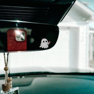 May include: A white ghost with a red heart in its hand is attached to the rearview mirror of a car. The ghost is a car charm.