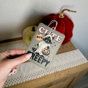 Op de afbeelding: Een grijze autoluchtverfrisser met een cartoonwasbeer die een geestkostuum draagt en een jack-o-lantern vasthoudt. De tekst op de luchtverfrisser luidt "Cute But Creepy".