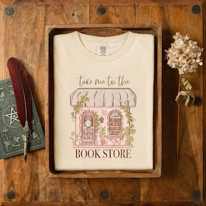 Pu&ograve; includere: Una maglietta bianca con una grafica di una libreria rosa e marrone con il testo "Take me to the Book Store".