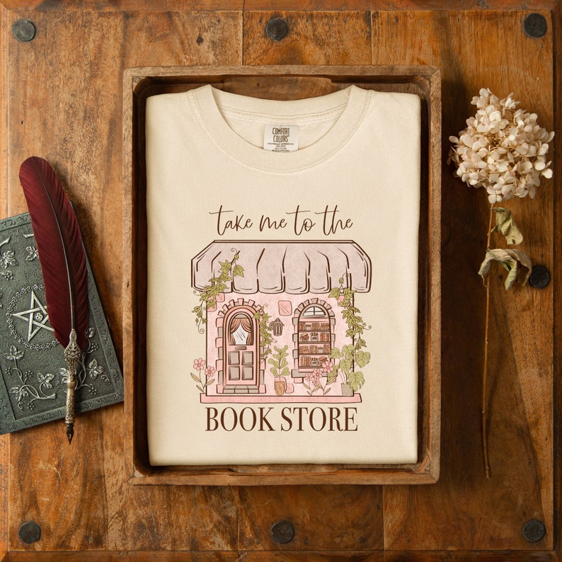 Pu&ograve; includere: Una maglietta bianca con una grafica di una libreria rosa e marrone con il testo "Take me to the Book Store".