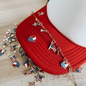 Puede incluir: Gorra de camionero roja, blanca y azul con una cadena dorada con colgantes de banderas americanas, estrellas y corazones. Hay colgantes adicionales de banderas y estrellas esparcidos alrededor de la gorra.