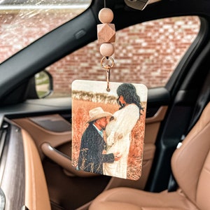 May include: Rectangular car air freshener with a photo of a couple kissing in a field. The air freshener is attached to a string with wooden beads and a gold-colored clip. The car interior is visible in the background.