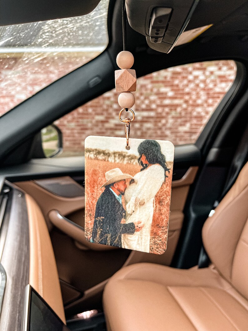 May include: Rectangular car air freshener with a photo of a couple kissing in a field. The air freshener is attached to a string with wooden beads and a gold-colored clip. The car interior is visible in the background.