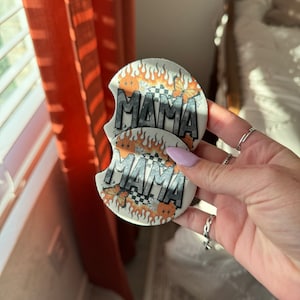 May include: Two white round coasters with a flame design and the word "MAMA" printed in black. The coasters have a checkered pattern and a butterfly design.