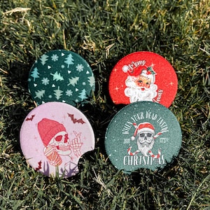 May include: Four round Christmas-themed coasters with different designs. One coaster features a green background with white snowflakes and green Christmas trees. Another coaster has a red background with a white Santa Claus. The third coaster has a pink background with a skull wearing a pink beanie and holding a cup. The fourth coaster has a green background with a skull wearing a Santa hat and the text "When you're dead inside, just say it's Christmas".