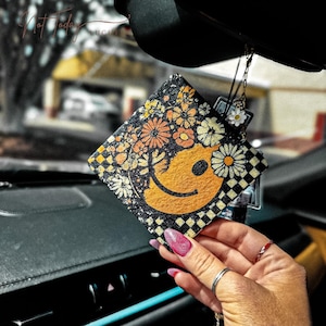 Puede incluir: Un ambientador de coche cuadrado con un diseño retro. Presenta una cara sonriente amarilla y un estampado floral en naranja, amarillo y blanco. El fondo es negro con un patrón de cuadros. El texto "Not Today" es visible.