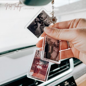 May include: Clear acrylic car charm with three rectangular photo inserts. The charm is suspended from a gold-coloured chain and features a daisy charm. The photos are black and white and depict people and buildings. The text "Not Today" and "LUCIIFER" are visible.
