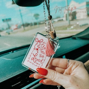 May include: A white car air freshener with a pink tassel hanging from it. The air freshener has a clear glass bottle and a white card with the text "You are so Loved" and "John 3:16" printed on it.