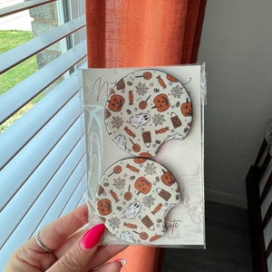 May include: Two white round beverage coasters with a Halloween theme. The coasters feature a pattern of orange pumpkins, ghosts, candy corn, spiderwebs, and stars on a white background.