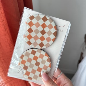 Puede incluir: Dos posavasos redondos para coche con un diseño a cuadros en tonos naranja, marrón y crema, empaquetados en plástico transparente. Los posavasos están diseñados para el portavasos del coche.
