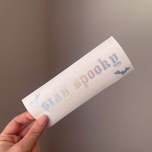 K&ouml;nnte beinhalten: Ein wei&szlig;er Aufkleber mit schimmerndem Regenbogen-Schriftzug, der "Stay Spooky" lautet, mit zwei schwarzen Fledermaus-Silhouetten auf jeder Seite.