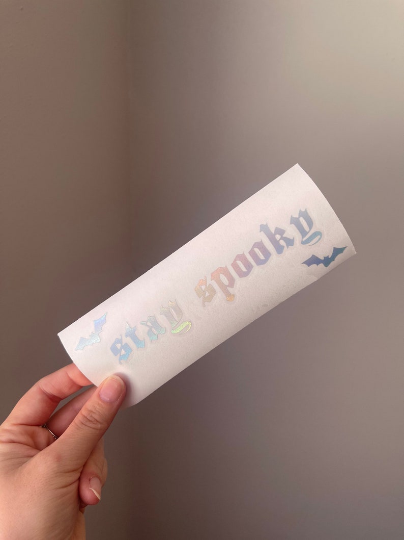 K&ouml;nnte beinhalten: Ein wei&szlig;er Aufkleber mit schimmerndem Regenbogen-Schriftzug, der "Stay Spooky" lautet, mit zwei schwarzen Fledermaus-Silhouetten auf jeder Seite.