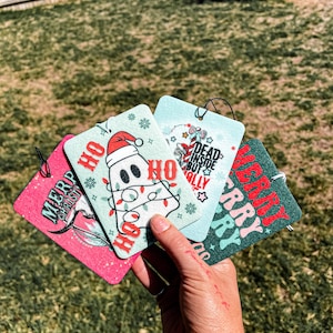 May include: Four Christmas-themed car air fresheners.  The air fresheners are green, pink, and white with various holiday designs and text.  The text on the air fresheners includes "Merry Christmas", "Ho Ho Ho", "Dead Inside But Festive", and "Merry & Bright".