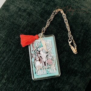 May include: A clear plastic charm with a turquoise background and a skull with flowers and the text "Last Night We Let The Liquor Talk". The charm is attached to a gold chain with a red tassel.