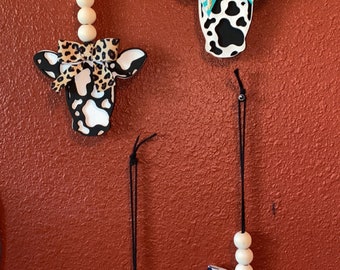 Puede incluir: Tres adornos de cabeza de vaca con estampado de vaca blanco y negro y un lazo. Los lazos son de diferentes patrones: estampado de leopardo, turquesa y blanco, y blanco y negro. Cada adorno tiene una cuerda de cuentas de madera y una etiqueta turquesa.