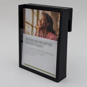 Puede incluir: Un portafolletos de plástico negro montado en la pared con un bolsillo frontal transparente. El soporte contiene una foto de una mujer y una tarjeta blanca con una franja verde. La mujer está mirando por una ventana.
