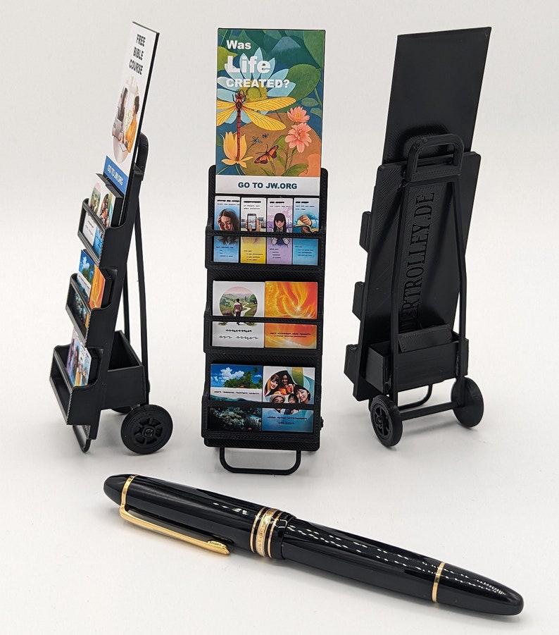 May include: A black pen with gold detailing lies in front of three black brochure display carts. The carts have wheels and shelves displaying cards with images and text. One cart reads "FREE BIBLE COURSE", another "Was Life Created?" and "GO TO JW.ORG".