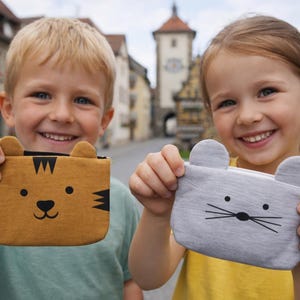 Könnte beinhalten: Zwei lächelnde Kinder halten kleine Beutel hoch. Einer ist braun mit einem Tigergesicht, der andere grau mit einem Mausgesicht. Die Beutel haben Reißverschlüsse und sind für kleine Gegenstände geeignet. Im Hintergrund ist eine Stadt zu sehen.
