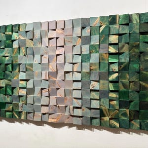 May include: A rectangular wooden wall art piece composed of numerous small, square blocks. The blocks transition in color from green to gray and tan, creating a gradient effect. The artwork is mounted on a white wall.