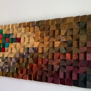 Puede incluir: Una pieza de arte de pared hecha de madera con un patrón geométrico. La madera está teñida en varios tonos de marrón, rojo, verde y azul. La pieza está montada en una pared blanca.
