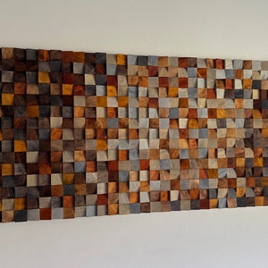 K&ouml;nnte beinhalten: Ein Wandkunstwerk aus Holz mit einem Mosaik aus verschiedenen farbigen Holzquadraten. Die Quadrate sind in einem Muster angeordnet, das einen wellenartigen Effekt erzeugt. Das Holz ist eine Mischung aus braunen, grauen und roten T&ouml;nen.