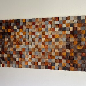 K&ouml;nnte beinhalten: Ein Wandkunstwerk aus Holzbl&ouml;cken in verschiedenen Braunt&ouml;nen, Graut&ouml;nen und Beiget&ouml;nen. Die Bl&ouml;cke sind in Wellenform angeordnet und schaffen ein einzigartiges und modernes Design.