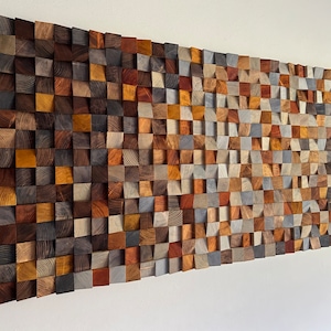 Könnte beinhalten: Ein großes, rechteckiges Wandkunstwerk aus Holz, das aus zahlreichen kleinen, quadratischen Blöcken besteht. Die Blöcke zeigen eine Vielzahl natürlicher Holztöne, darunter Braun-, Grau- und Orangetöne, wodurch ein strukturiertes, mosaikartiges Aussehen entsteht. Das Kunstwerk ist an einer weißen Wand befestigt.