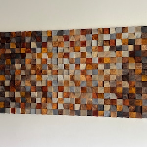K&ouml;nnte beinhalten: Ein Wandkunstwerk aus Holzquadraten in verschiedenen Braunt&ouml;nen, Graut&ouml;nen und Beiget&ouml;nen. Die Quadrate sind in Wellenform angeordnet und schaffen ein einzigartiges und modernes Design.