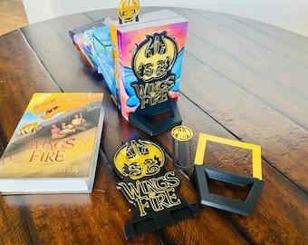 May include: A black bookend with a gold dragon design and the words "Wings of Fire" on it. There are several books stacked behind the bookend, and a smaller bookend with the same design is in the foreground.