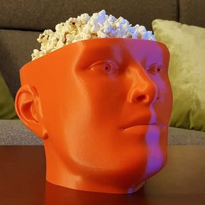 May include: An orange, human-head-shaped container filled with popcorn. The container is a solid, smooth orange color, with facial features including eyes, a nose, and lips. The popcorn is white and fluffy, overflowing from the top of the container.