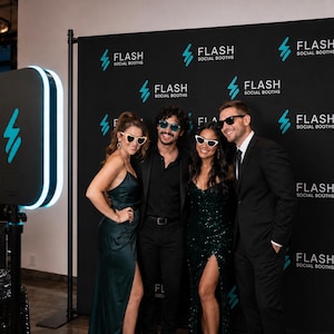 May include: A portable photo booth kiosk with a glowing blue light ring stands next to a black step-and-repeat backdrop featuring the white text FLASH SOCIAL BOOTHS. A small sign reads GRAB A PROP STRIKE A POSE SHARE THE LOVE near various novelty glasses.