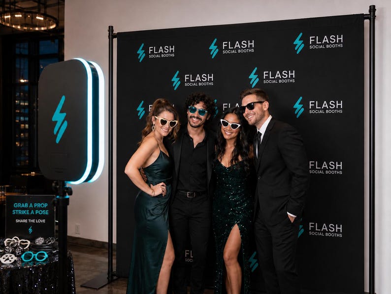 May include: A portable photo booth kiosk with a glowing blue light ring stands next to a black step-and-repeat backdrop featuring the white text FLASH SOCIAL BOOTHS. A small sign reads GRAB A PROP STRIKE A POSE SHARE THE LOVE near various novelty glasses.