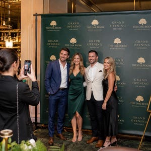 May include: A dark green step and repeat backdrop featuring the gold text "THE OAK ROOM KITCHEN & COCKTAILS" and "GRAND OPENING VIP EVENT" in a repeating pattern. A matching sign on an easel stands to the right with the same branding and text.