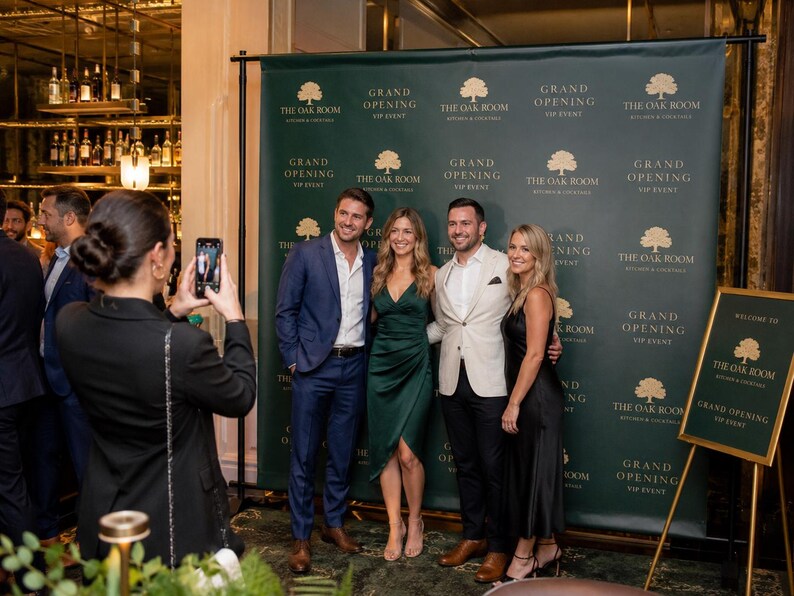 May include: A dark green step and repeat backdrop featuring the gold text "THE OAK ROOM KITCHEN & COCKTAILS" and "GRAND OPENING VIP EVENT" in a repeating pattern. A matching sign on an easel stands to the right with the same branding and text.