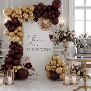 Pu&ograve; includere: Allestimento elegante per eventi con palloncini dorati e bordeaux che formano un arco. Un cartello recita "Love is Brewing". Macchina da caff&egrave;, composizioni floreali e candele completano la scena.