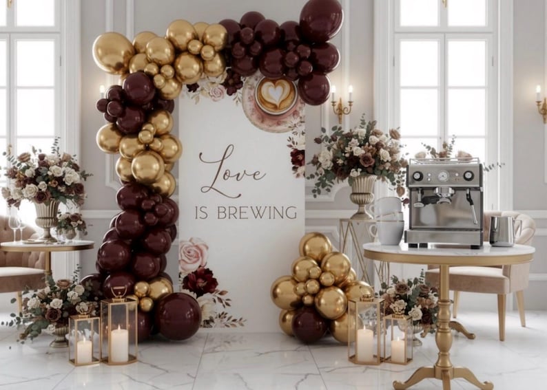 Pu&ograve; includere: Allestimento elegante per eventi con palloncini dorati e bordeaux che formano un arco. Un cartello recita "Love is Brewing". Macchina da caff&egrave;, composizioni floreali e candele completano la scena.