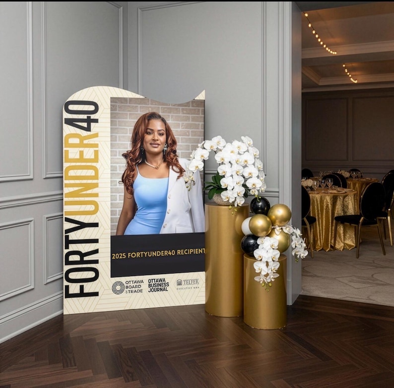 Pu&ograve; includere: Un grande display con la scritta "FORTYUNDER40" e una foto di una donna con un abito blu e un blazer bianco. Due piedistalli cilindrici dorati sostengono orchidee bianche e palloncini neri e dorati. L'evento si svolge in una sala da ballo.
