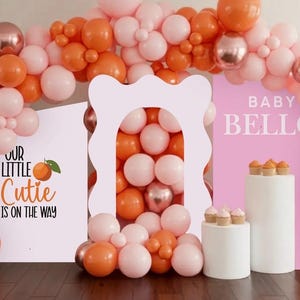 Pu&ograve; includere: Sfondo per baby shower con arco di palloncini rosa, arancioni e oro rosa. Cartelli con la scritta "Our Little Cutie is on the Way" e "Baby Bello". Piedistalli bianchi con cupcakes.
