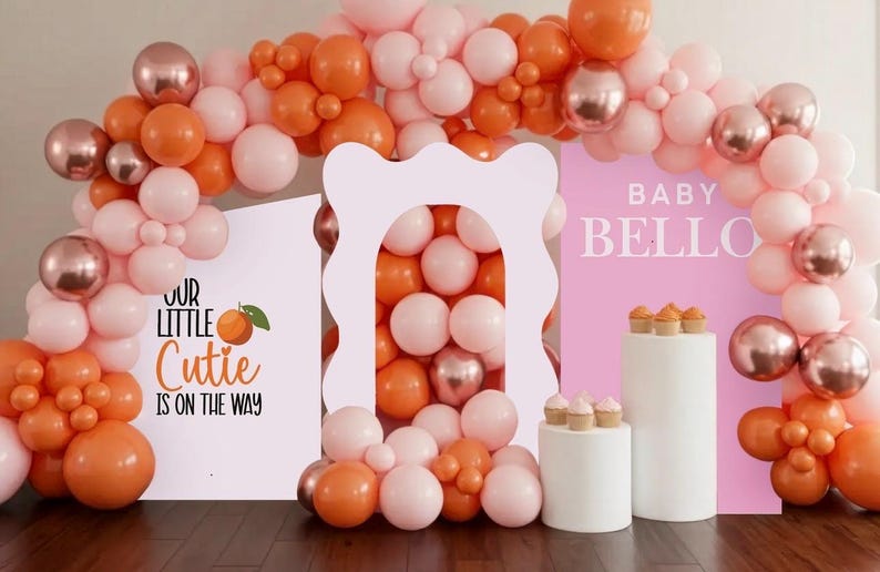 Pu&ograve; includere: Sfondo per baby shower con arco di palloncini rosa, arancioni e oro rosa. Cartelli con la scritta "Our Little Cutie is on the Way" e "Baby Bello". Piedistalli bianchi con cupcakes.