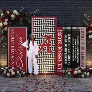 Pu&ograve; includere: Una scena di celebrazione della laurea con una donna in completo bianco che posa davanti a pannelli decorativi. I pannelli presentano il logo dell'Universit&agrave; dell'Alabama, la scritta "Congratulazioni Asiriah" e "Classe del 2025". Composizioni floreali rosse e bianche incorniciano la scena.