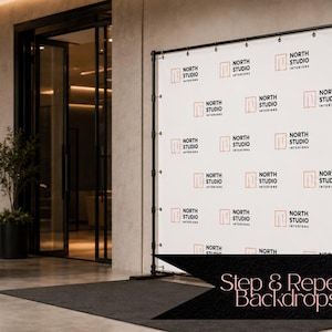 May include: A white step and repeat backdrop banner featuring a repeating pattern of the black text North Studio Interiors and an orange square logo. The banner is mounted on a black metal frame stand with grommets along the top edge.