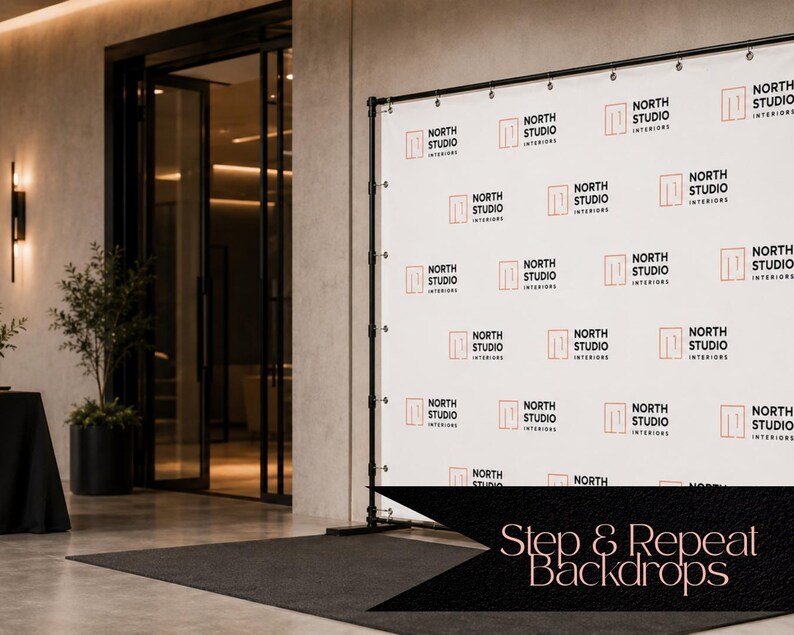 May include: A white step and repeat backdrop banner featuring a repeating pattern of the black text North Studio Interiors and an orange square logo. The banner is mounted on a black metal frame stand with grommets along the top edge.