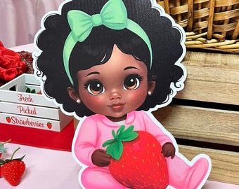 May include: A die-cut image of a baby girl with dark skin, wearing a pink outfit and holding a large red strawberry. She has black curly hair, a green bow, and a green headband. The image is on a pink surface.