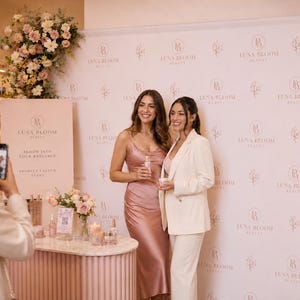 May include: A promotional event backdrop featuring the repeating Luna Bloom Beauty logo and floral illustrations in rose gold. Two individuals stand in front of the banner, one in a satin pink midi dress and one in a cream-colored blazer and trousers.