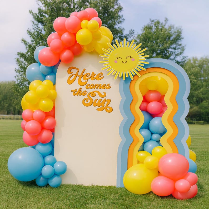 Pu&ograve; includere: Un fondale fotografico decorativo con la scritta "Here comes the Sun" e un ritaglio di sole sorridente. Lo sfondo &egrave; decorato con palloncini colorati nei toni del rosa, giallo e blu, creando un'atmosfera allegra e festosa.